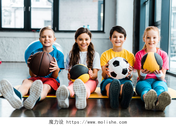 在灰色背景教室里面有四个青春期的小孩手里拿着不同的健身球青春期前的孩子坐在健身垫与球